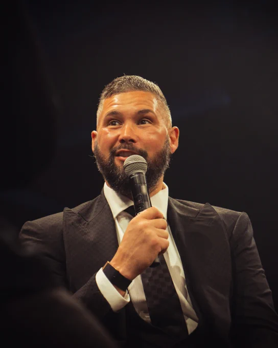 A man in a suit speaks into a microphone at a formal event. The lighting is warm, creating an engaging atmosphere.