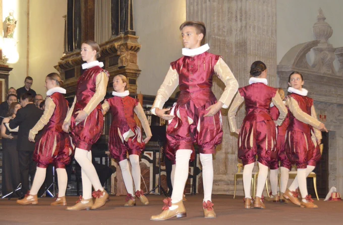 Dansa dels Infantillos del Corpus de València. Grupo de jóvenes bailando en escenario con trajes renacentistas burdeos y medias blancas, en interior tipo iglesia.