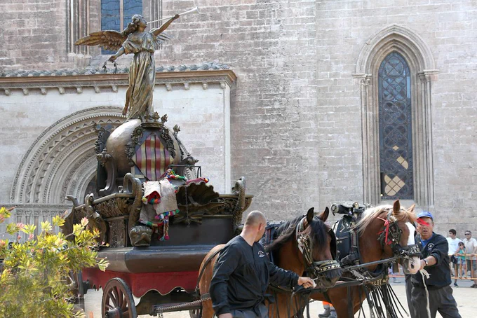 Roca de La Fama del Corpus de València. Carroza ornamentada con estatua alada y escudo, tirada por dos caballos guiados por hombres frente a una iglesia gótica.