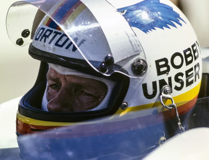 A close up of Bobby Unser in the cockpit, wearing his crash helmet with the visor raised and looking focused. His crash helmet is white with his name painted in black capital letters on the side, the head of a blue eagle and yellow, orange, red and blue stripes around the base and over the top. 1981 CART Phoenix.
