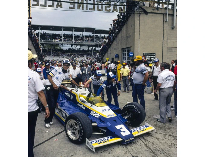 Penske team members push the blue, white and yellow Penske PC9B through the passageway between two grandstands into Gasoline Alley, in the infield of the track. The area is bustling with people, with the large, tiered grandstand on the other side of the track visible through the passageway.