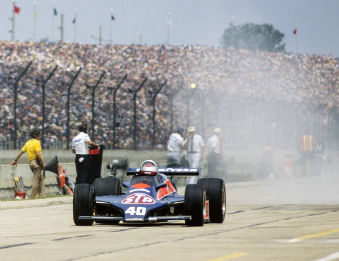 Mario Andretti heads along pit road in his blue and red, STP sponsored Patrick Racing Wildcat Mk8. Tire smoke clouds the view behind him, with various team members standing along pit wall, looking the other way. In the background, the huge grandstands are completely filled with people.