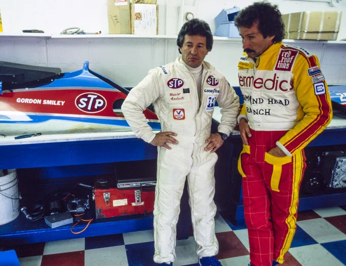 On the right of frame, Mario Andretti stands wearing a white fire suit with his hands on his hips; beside him on the far right stands Gordon Smiley, wearing a red, yellow and white fire suit with his hands in his pockets. They are in Smiley's garage area, with blue and red bodywork from Smiley's car seen on top of blue cabinets behind them.