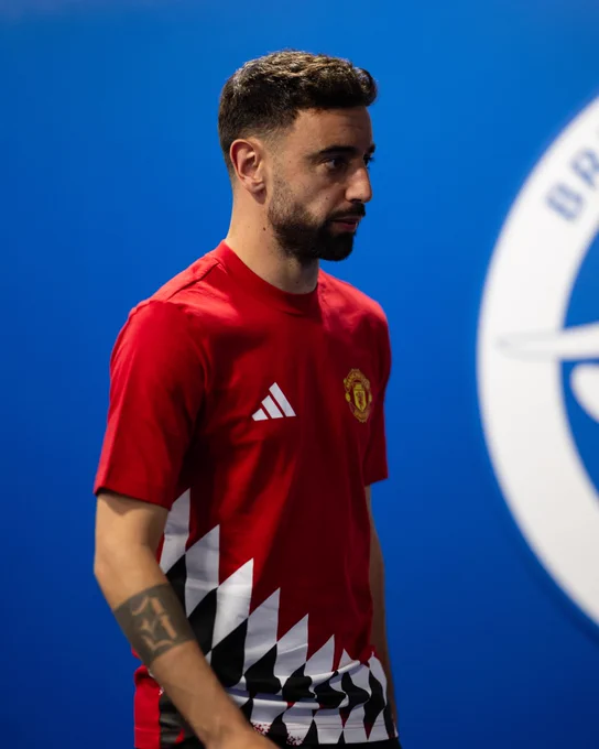 Bruno Fernandes wearing a red Manchester United shirt with a geometric black and white pattern on the side, standing against a blue background.