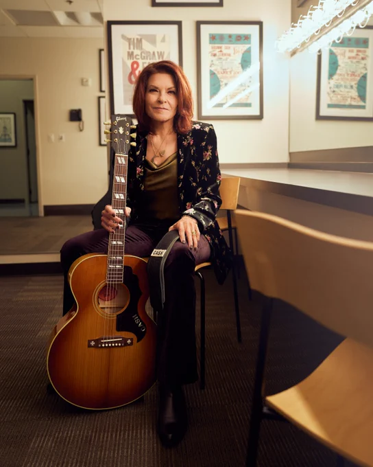 Rosanne Cash is seated in a dressing room holding her Gibson signature acoustic guitar. The room has framed posters on the walls. The atmosphere appears calm and focused.