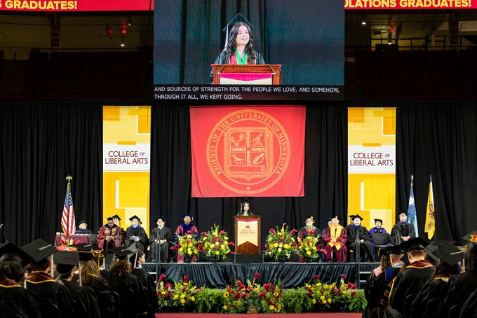 A female speaker in graduation attire delivers a speech on stage at commencement with faculty seated behind her.