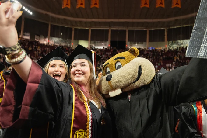 Two graduates in caps and gowns take a selfie with Goldy inside a crowded indoor arena.