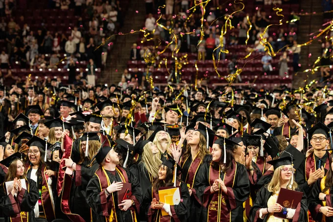 Graduates in caps and gowns celebrating by tossing their caps and releasing gold streamers in a crowded auditorium.