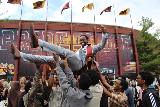 Group of students joyfully lifting a graduate wearing a maroon and gold stole in front of Mariucci.