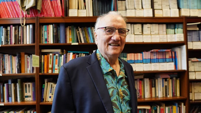 Walt Wolfram wearing a dark blazer and patterned shirt standing in front of a large bookshelf filled with books and files.