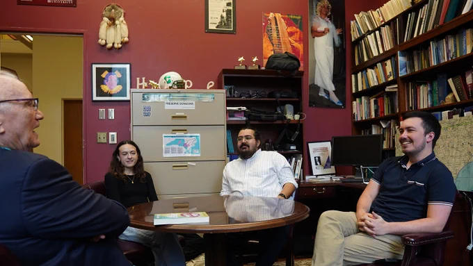 Four people sitting around a round wooden table in a cozy office filled with bookshelves and framed artwork on the walls.