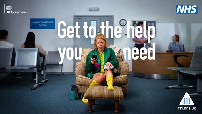 A woman wearing sportswear is sat on an armchair. The woman is nursing an injury to her leg whilst using a phone in her other hand. The armchair is placed in the middle of an urgent treatment centre waiting room.