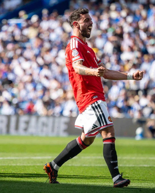 Bruno Fernandes in a red jersey and black socks celebrating on the field with a crowd in the background.