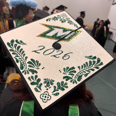 A decorated graduation cap with "2026" and a green and gold emblem with the letter M surrounded by green glittery floral designs. Graduates in caps and gowns are in the background.