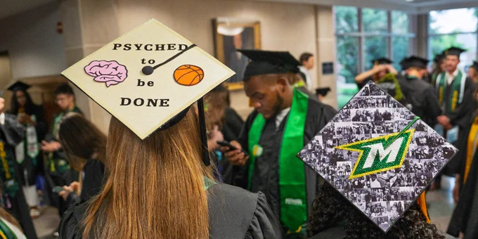 Students at a graduation ceremony are wearing caps and gowns. One cap has a design with a brain, basketball, and the words "Psyched to be done." Another cap features a collage with the letter "M."