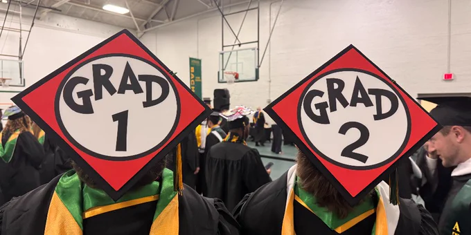 Graduates in caps and gowns facing forward, with the words "Grad 1" and "Grad 2" on their caps in a gymnasium setting.