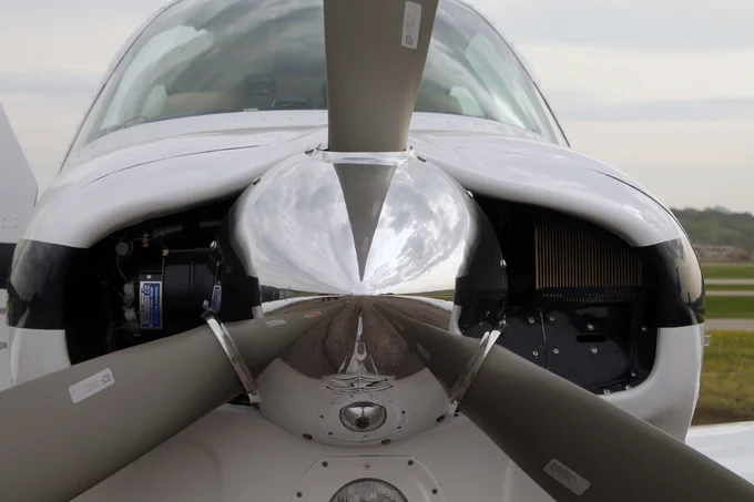 Close-up of a Beechcraft Bonanza aircraft's silver propeller, with reflections visible on its polished surface. The engine area is partially exposed on both sides. The background shows a cloudy sky and grassy terrain.