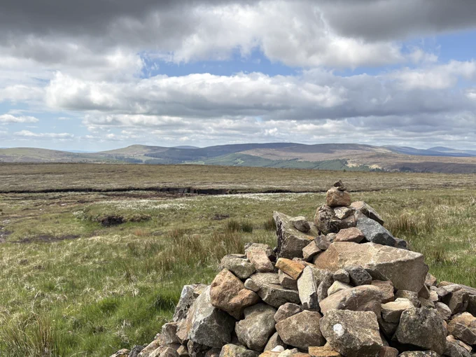 A landscape photograph. A stone cairn sits to the right foreground of a grassy landscape strewn with white bog cotton flowers. This leads to a valley, from which a mountain range rises in the far distance. The sky is blue, but there is a covering of puffy white clouds.