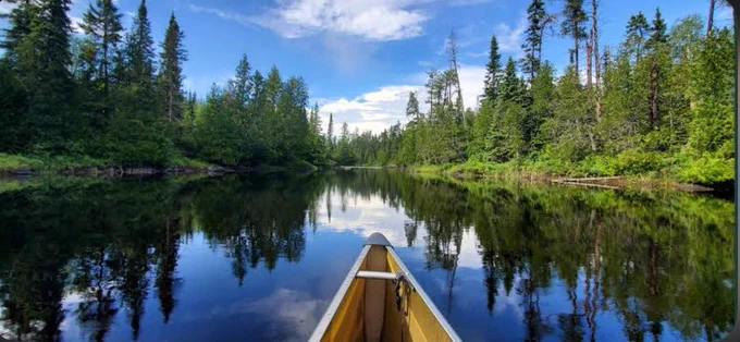 A canoe moves through the calm waters of Boundary Waters Lake in Minnesota, surrounded by dense evergreen forest beneath a bright blue sky reflected like a mirror across the pristine wilderness waterway.