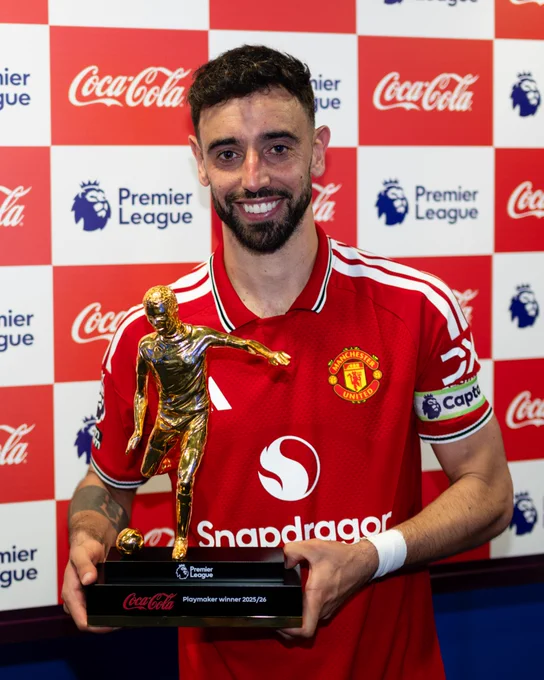 Bruno Fernandes wearing his captain's armband holds the Premier League Playmaker award trophy in front of Coca-Cola and Premier League backdrop.