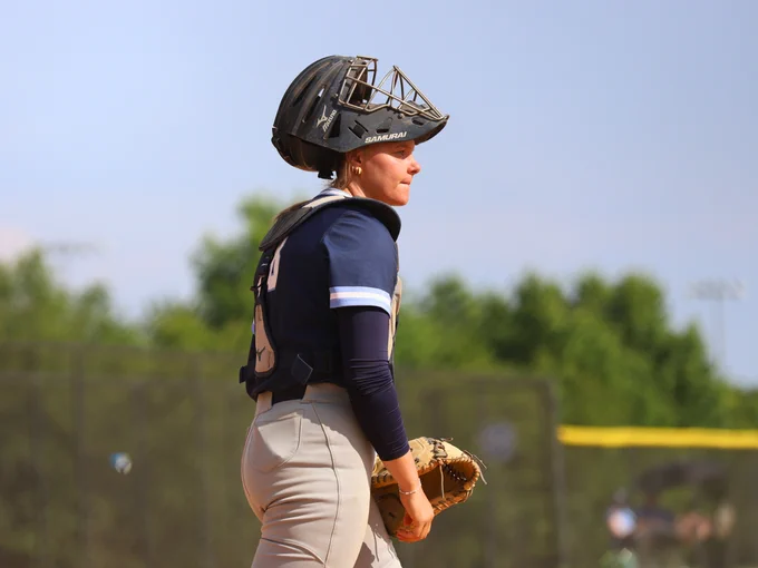 Softball catcher wearing full gear on a sunny day with a blurred green field background.