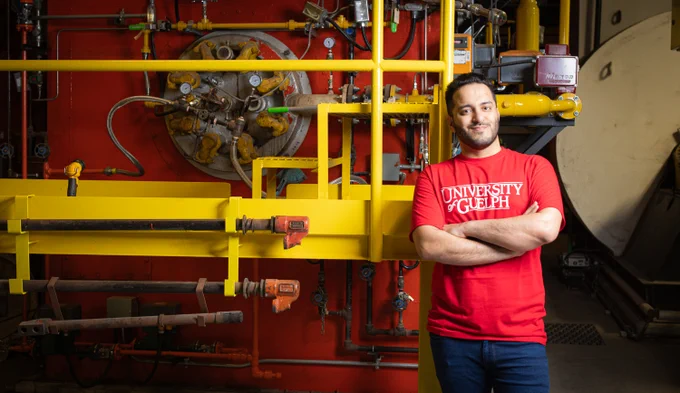 Shahriyar Ghazanfari Holagh, a 2025 PhD in Mechanical Engineering, stands in front of a yellow machine. He is wearing a red University of Guelph t-shirt. His arms are crossed and he has a soft smile. 