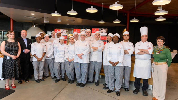 A group of chefs and individuals stand together in a kitchen setting, with some wearing chef uniforms and hats. They are posing in front of a counter, with cooking-related materials visible in the background.