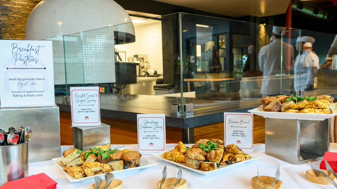 An assortment of breakfast pastries is displayed on a table with several trays. A sign mentions "Breakfast Pastries made by graduate learners of Montco's Culinary Institute.
