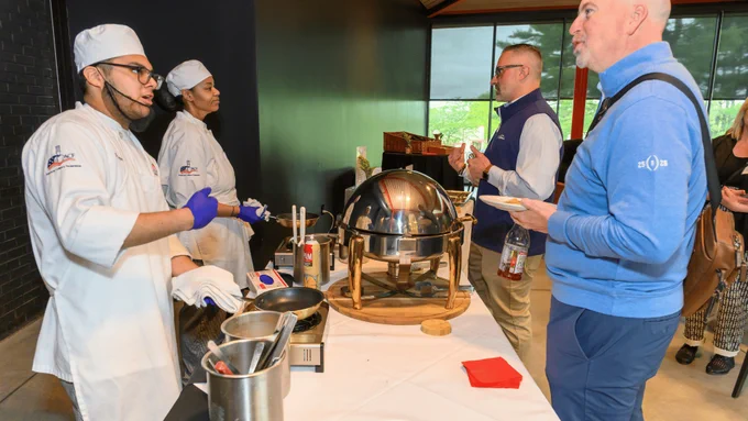 A chef in a white uniform serves food from a buffet setup at a culinary event. Another person in similar attire stands nearby. Guests holding plates engage in conversation. The setting appears to be indoors with large windows offering a view of greenery outside.