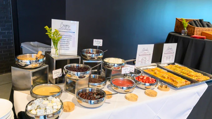 A buffet table features a crepe station with various toppings and ingredients. There are bowls of chocolate chips, strawberries, whipped cream, and sauces. The setup includes decorative greenery and a dark tablecloth in the background.