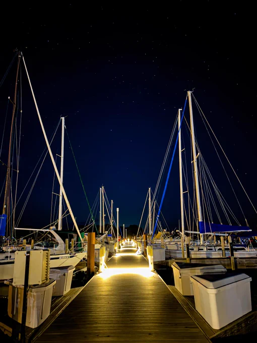 Nighttime view of a marina with several docked sailboats under a starry sky, illuminated by lights along the dock.