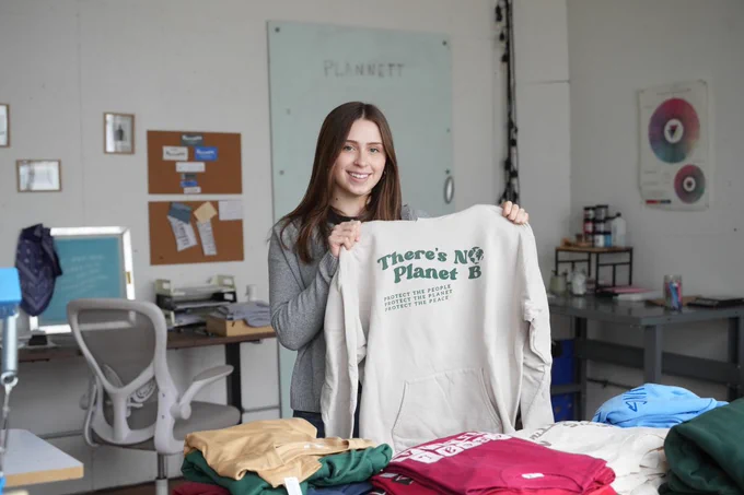 WMU student Julia Adams is smiling and holding a hoodie with the text "There's No Planet B" printed on it. The room features a desk with a computer, a chair, and various colorful garments folded on a table. A board with notes and posters is visible in the background.