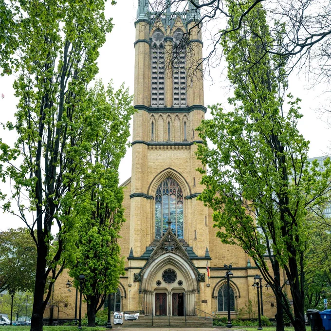 Tall Gothic-style church tower with pointed spires framed by green leafy trees on a cloudy day.