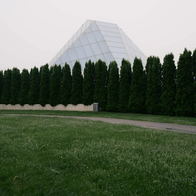 Modern glass pyramid structure partially hidden behind a tall row of dense evergreen trees on a cloudy day.