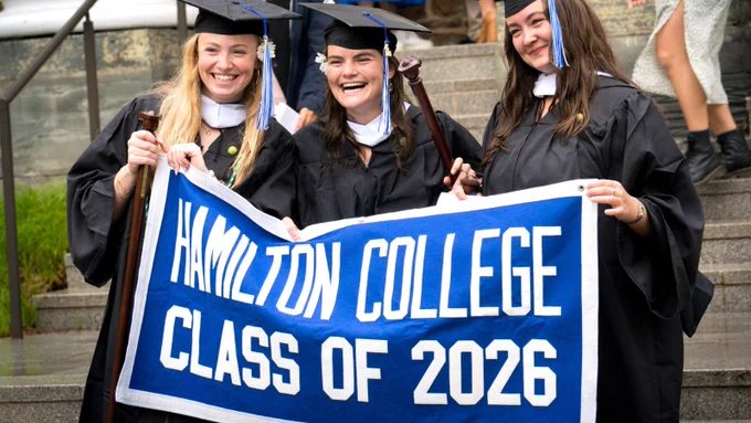 Three graduates in caps and gowns smile while holding a banner that reads, "Hamilton College Class of 2026." They are standing on outdoor steps.