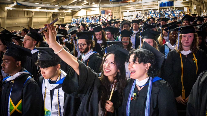 A group of graduates in caps and gowns gather for a selfie during a commencement ceremony. The venue is filled with people, and there's a visible Jamaican flag on one gown. The atmosphere is celebratory.