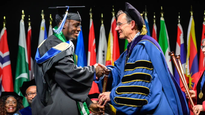 Two individuals in academic regalia are shaking hands on a graduation stage, with international flags in the background.