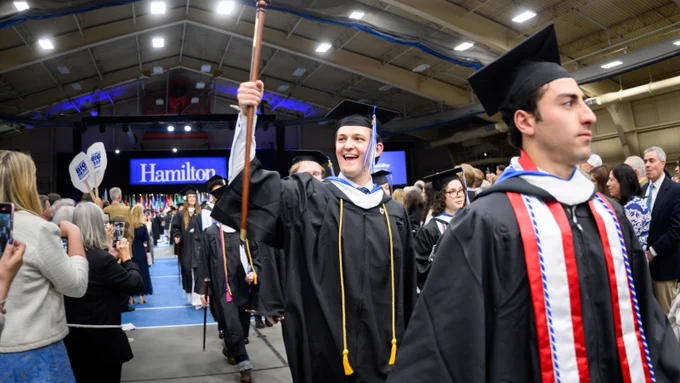 Graduates in academic regalia walk down an aisle at a commencement ceremony. One holds a ceremonial staff while smiling. In the background, there's a sign with "Hamilton" displayed.