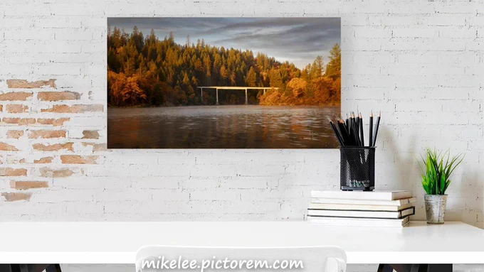 The historic Cottonwood Creek railroad trestle spans above Lake Britton in Shasta County, California, on an autumn morning. The steel and timber structure rises above the calm lake surface, its weathered ironwork framed by shoreline trees in full fall color. Warm morning light breaks over the eastern ridge and catches the turning foliage along the water's edge — yellows, oranges, and golds reflected in the still lake below. The adjacent campground is empty and the scene is quiet. The rails are gone now, the trestle a relic, recognizable to anyone who watched a group of twelve-year-olds run across it in 1986 and has carried that image ever since.