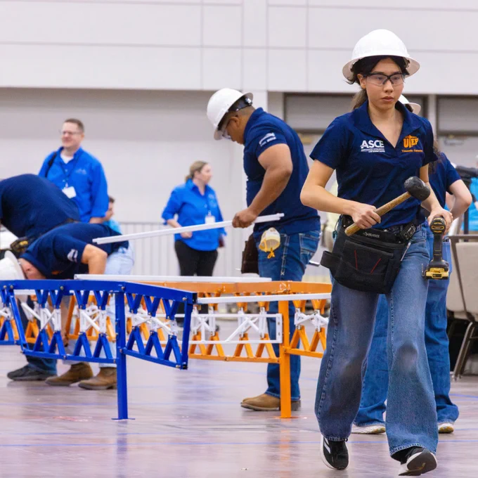 A member of the University of Texas at El Paso's student steel bridge team wearing  a blue shirt and hard hat walks carrying tools in a large convention center hall, with several others in similar attire working in the background. The setting features an ASCE and UTEP logo.