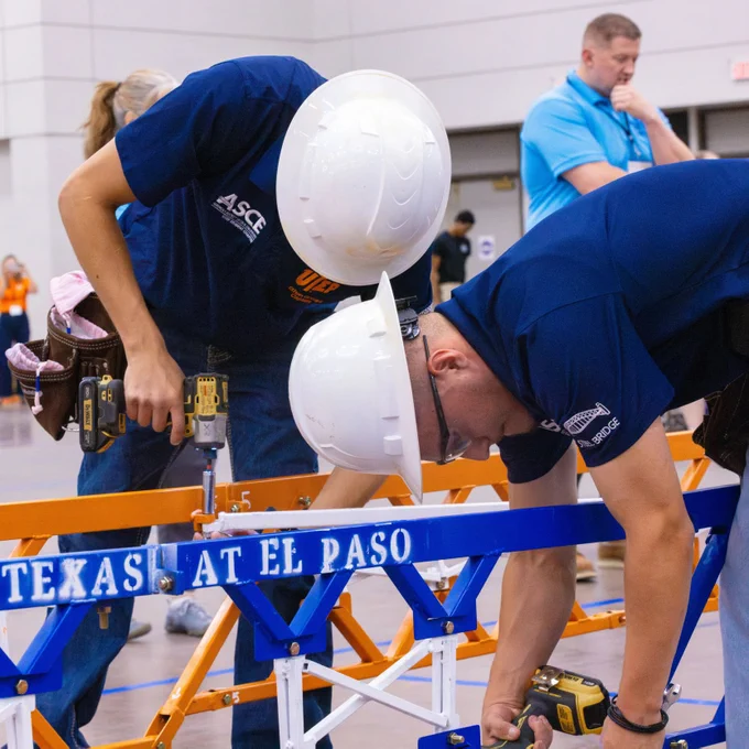 Two members of UTEP's student steel bridge team wearing hard hats and blue shirts are assembling a structure featuring the words "University of Texas at El Paso." They are focused on their task, using tools to secure components. A person in the background is observing the activity.