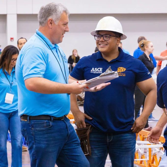 Two people are standing and conversing inside a convention center hall. One holds a clipboard and is wearing a blue polo shirt, while the other, a member of The University of Texas at El Paso's student steel bridge team, wears a hard hat and a shirt with the ASCE and UTEP logos. Other individuals are visible in the background.