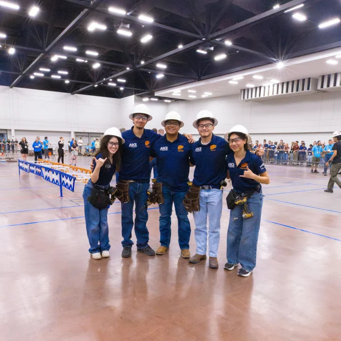 Members of The University of Texas at El Paso's student steel bridge team stand together indoors, wearing matching shirts and helmets. They are smiling and posing for a photo in a large hall. In the background, there are banners and spectators observing an event.
