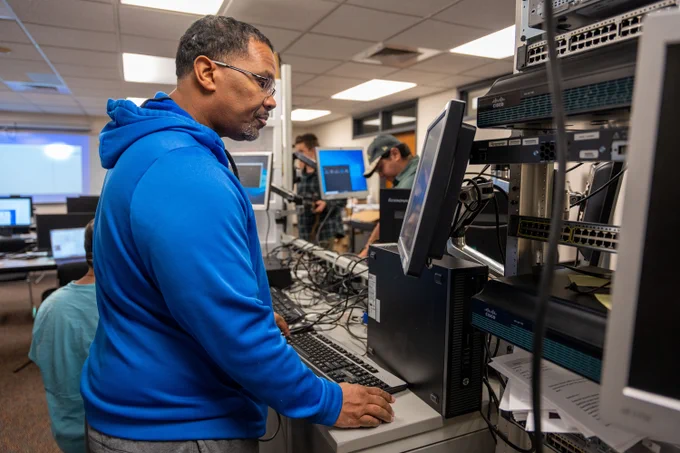 Person in blue hoodie working on a desktop computer in a busy tech lab filled with networking equipment and monitors.