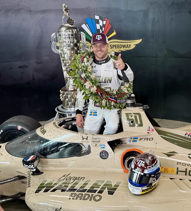 A person in a racing suit and cap stands on a race car, giving a thumbs-up gesture. A large trophy is behind them, and the Indianapolis Motor Speedway logo is visible. The race car displays "Morgan Wallen Radio" and various sponsor logos.