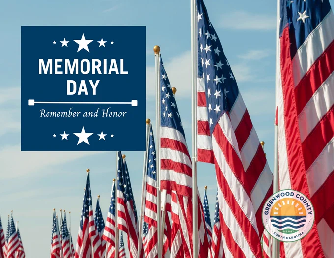 American flags are lined up under a blue sky. A sign reads, "Memorial Day Remember and Honor." The Greenwood County logo is visible in the corner.