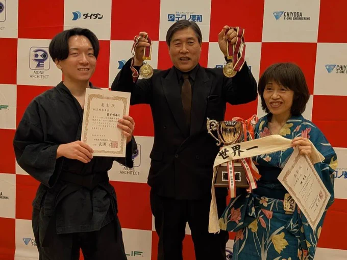 第五回世界棒手裏剣競技大会（神田明神ホール）にて、賞状を持つ剣王戦準優勝の亀井さん（左）、両手にメダルを掲げる和伝流手裏剣道の木﨑克彦宗家（中央）、世界一のタスキが巻かれたトロフィーと賞状を抱えて笑顔を見せる女王戦優勝の翠風さん（右）の記念3ショット。