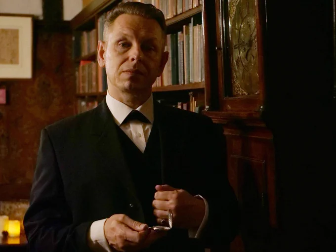 A smartly dressed man stands near a bookcase. There is a Grandfather clock next to him.