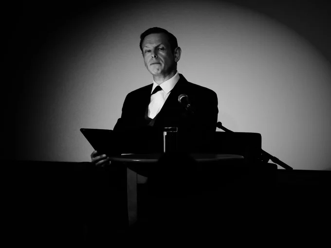 A man sits at a desk reading from a tablet. There is a microphone next to him.