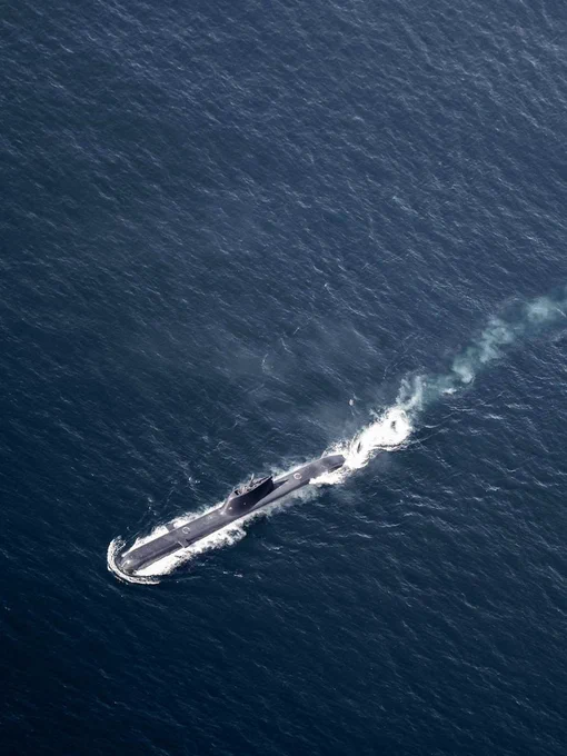 A military submarine cruising on the surface of open blue water, leaving a white wake and a faint trail of exhaust behind it. Photo by Standing NATO Maritime Group 1. 